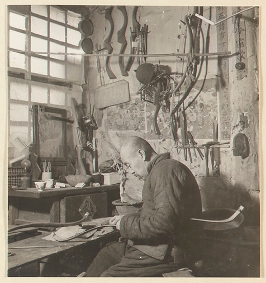 A man constructing a short monkey bow, some pellet crossbows can be seen on the wall behind him. Photo by Hedda Morrison at Ju Yuan Hao, Beijing, 1935.